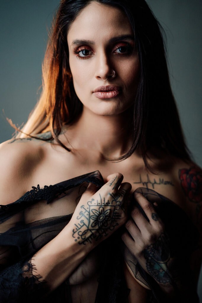 Una mujer de pelo castaño largo y tatuajes visibles en las manos y el pecho sujeta una tela negra transparente contra su pecho. Tiene un aro en la nariz, los ojos maquillados y una expresión neutra, con una iluminación suave que resalta sus rasgos sobre un fondo oscuro.