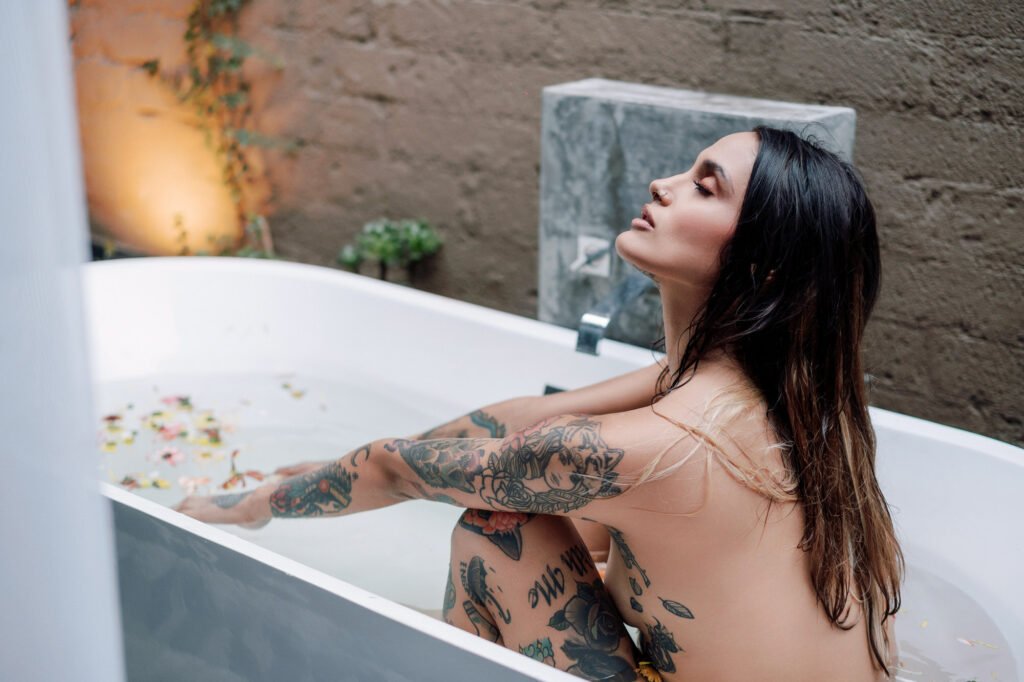 Una mujer tatuada con el pelo mojado está sentada desnuda en una bañera blanca llena de agua y pétalos de flores. Apoya los brazos en las rodillas e inclina la cabeza hacia atrás con los ojos cerrados, con aspecto relajado. El fondo muestra una pared de piedra y una iluminación interior ambiental.