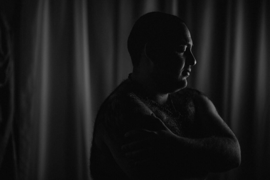 Una persona con la cabeza rapada se muestra de perfil con una dramática iluminación en blanco y negro, de pie contra unas cortinas suaves y desenfocadas. Sus brazos están cruzados sobre el pecho y su rostro está parcialmente iluminado, lo que acentúa el estado de ánimo contemplativo.