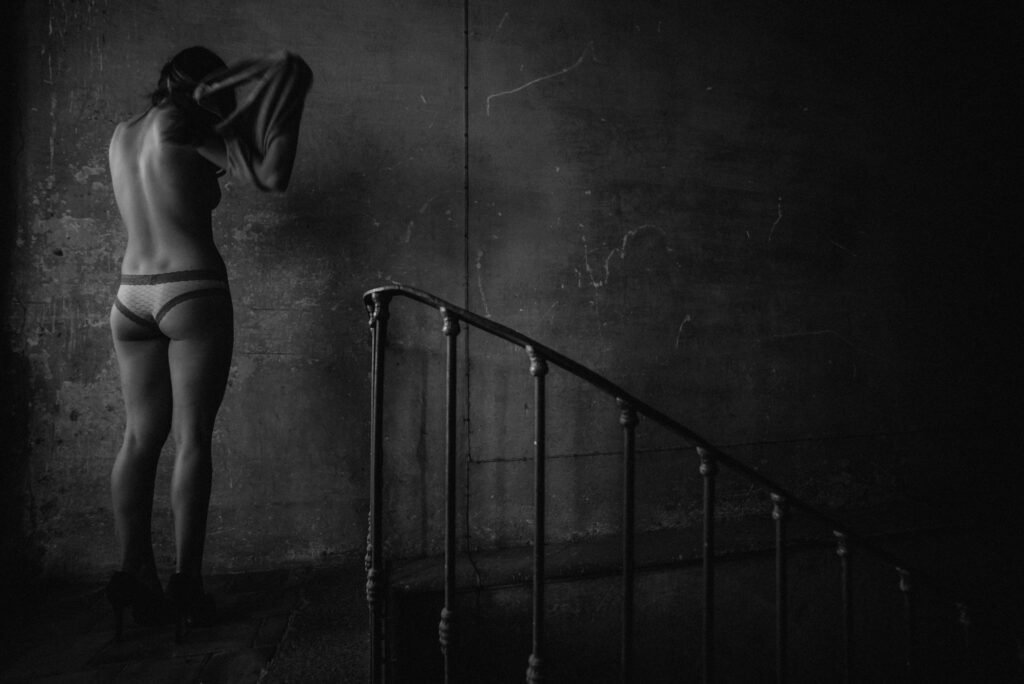 Una foto en escala de grises muestra a una persona en ropa interior y tacones, frente a una pared con textura y escasamente iluminada. Se levanta una camisa por encima de la cabeza, ocultando parcialmente su rostro. Una barandilla de hierro de la escalera se curva en primer plano, añadiendo profundidad a la escena sombría.