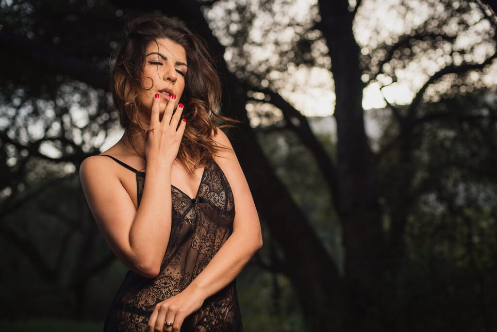 Una mujer con un vestido de encaje negro transparente está de pie al aire libre frente a unos árboles borrosos, tocándose la cara con la mano y cerrando los ojos. Lleva el pelo castaño despeinado y las uñas pintadas de rojo. La iluminación crea una atmósfera dramática y taciturna.