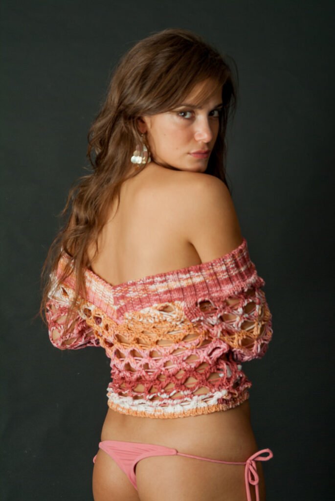 Una mujer posa delante de un fondo oscuro, mirando por encima del hombro. Lleva un top de crochet rosa y naranja sin hombros y una braguita de bikini rosa, con el pelo largo y castaño, pendientes de aro y una expresión facial seria.