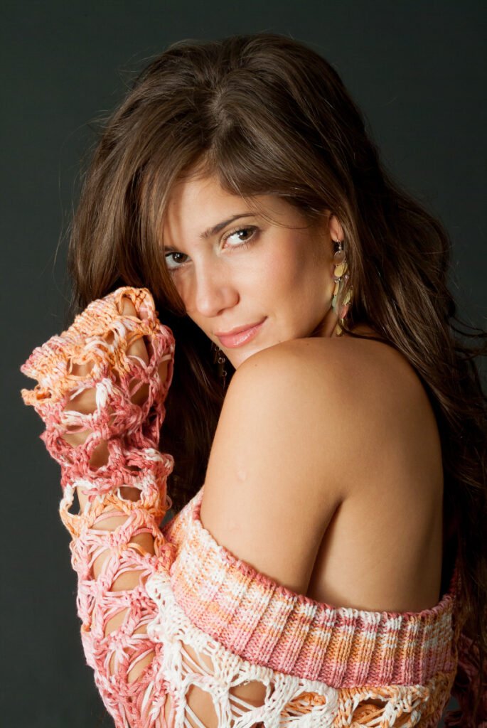 Una mujer de pelo largo y castaño posa sobre un fondo oscuro, mirando a la cámara por encima del hombro. Lleva un colorido jersey de ganchillo sin hombros en tonos rosa, naranja y blanco, y unos pendientes dorados.