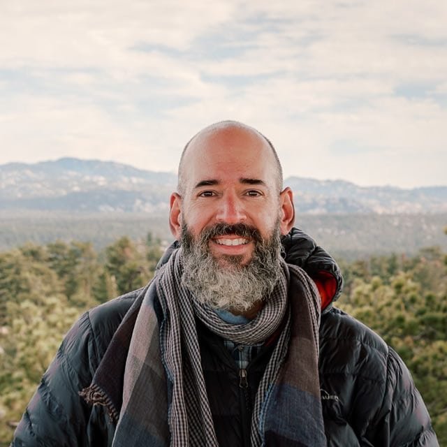Un hombre con barba y calvo sonríe a la cámara. Lleva una chaqueta oscura y una bufanda gris estampada. En el fondo se ven pinos y montañas lejanas bajo un cielo nublado.